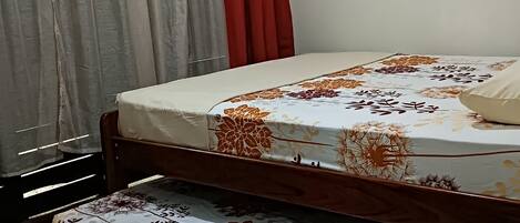 A room with a bed, a wooden floor, a window with curtains, and a view of greenery outside.