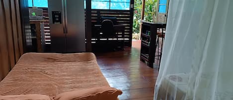 A room with a bed, a refrigerator, and a window with a view of greenery.