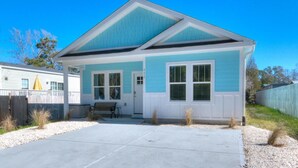 A single-story house with a light blue exterior and white trim. It features a front porch with a bench, a driveway, and a small yard with sparse vegetation. - Captain Jacks: Cozy 3BR Oak Island retreat, perfect for family fun and relaxation. (Oak Island)