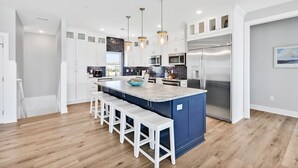 A modern kitchen with white cabinets, a blue island with bar stools, stainless steel appliances, and pendant lighting. - Experience beachfront luxury at Seaclusion, Oak Island's 10-bedroom retreat. (Oak Island)
