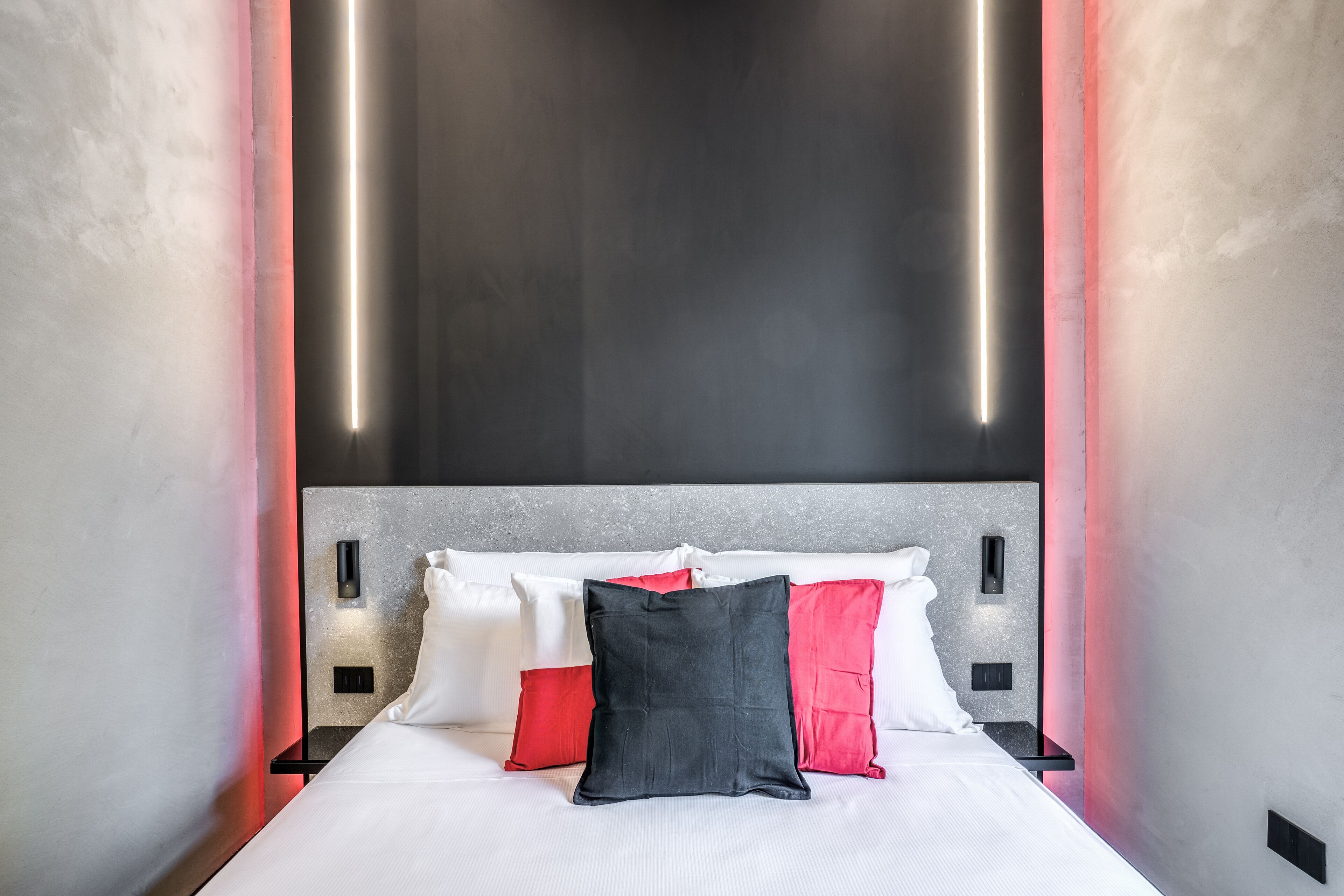 A modern hotel room with a large bed, white and red pillows, and a minimalist headboard.