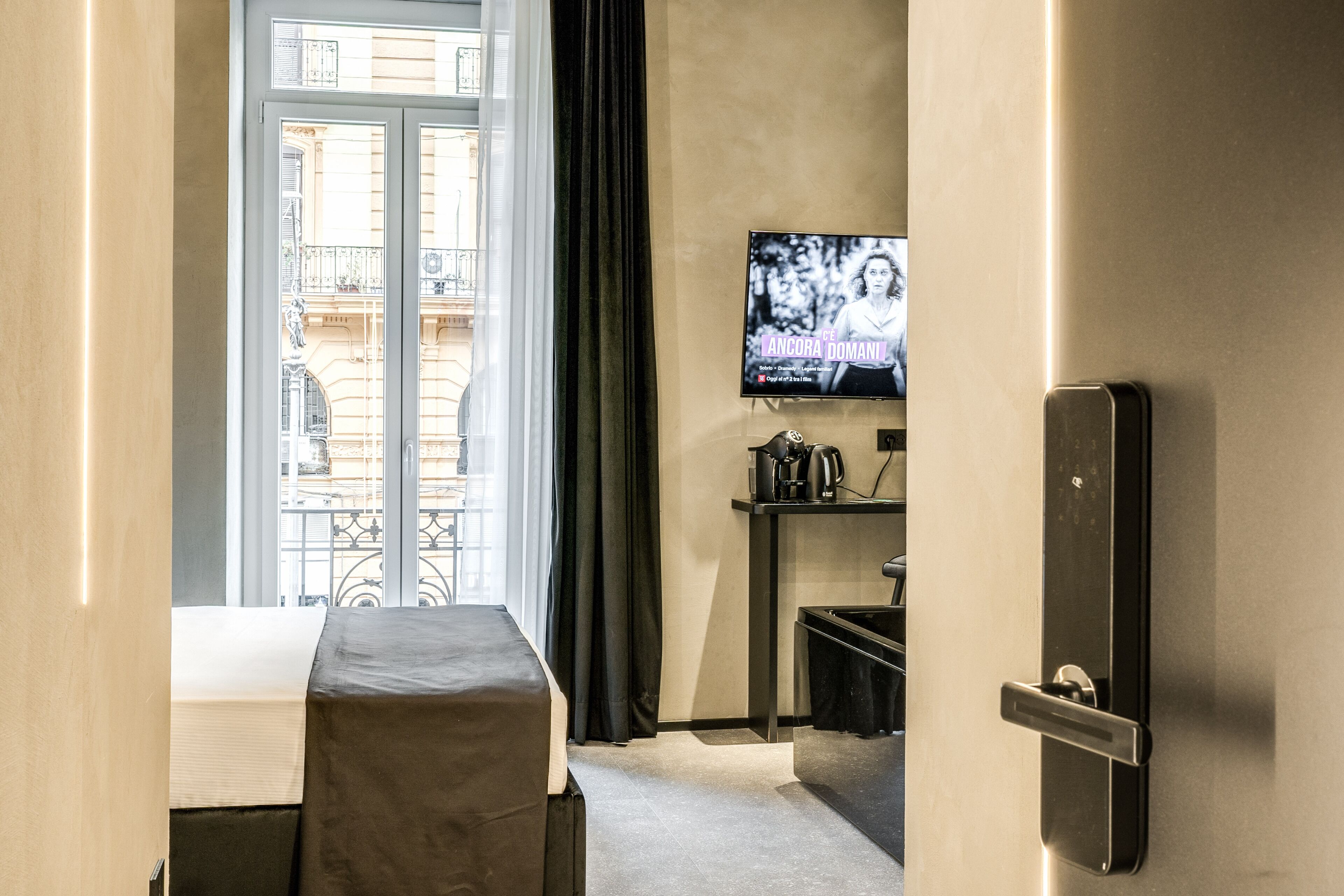 A hotel room with a bed, a television mounted on the wall, a window with curtains, and a view of the cityscape.