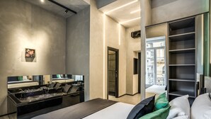 A modern hotel room with a large bed, a vanity with a mirror, a black console table, and a view of the city through a balcony door. - Gallery Experience Napoli (Naples)