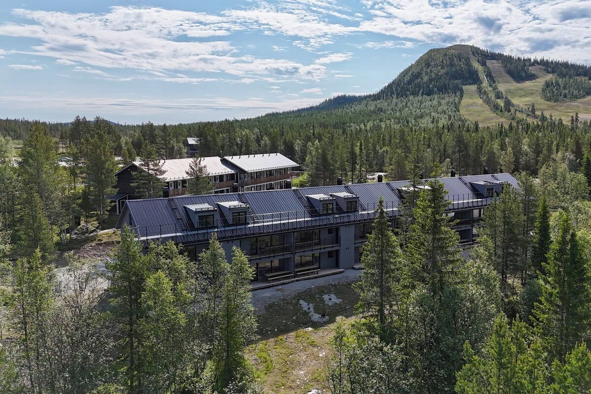 A modern building with a metal roof, surrounded by dense forest and a mountain in the background.