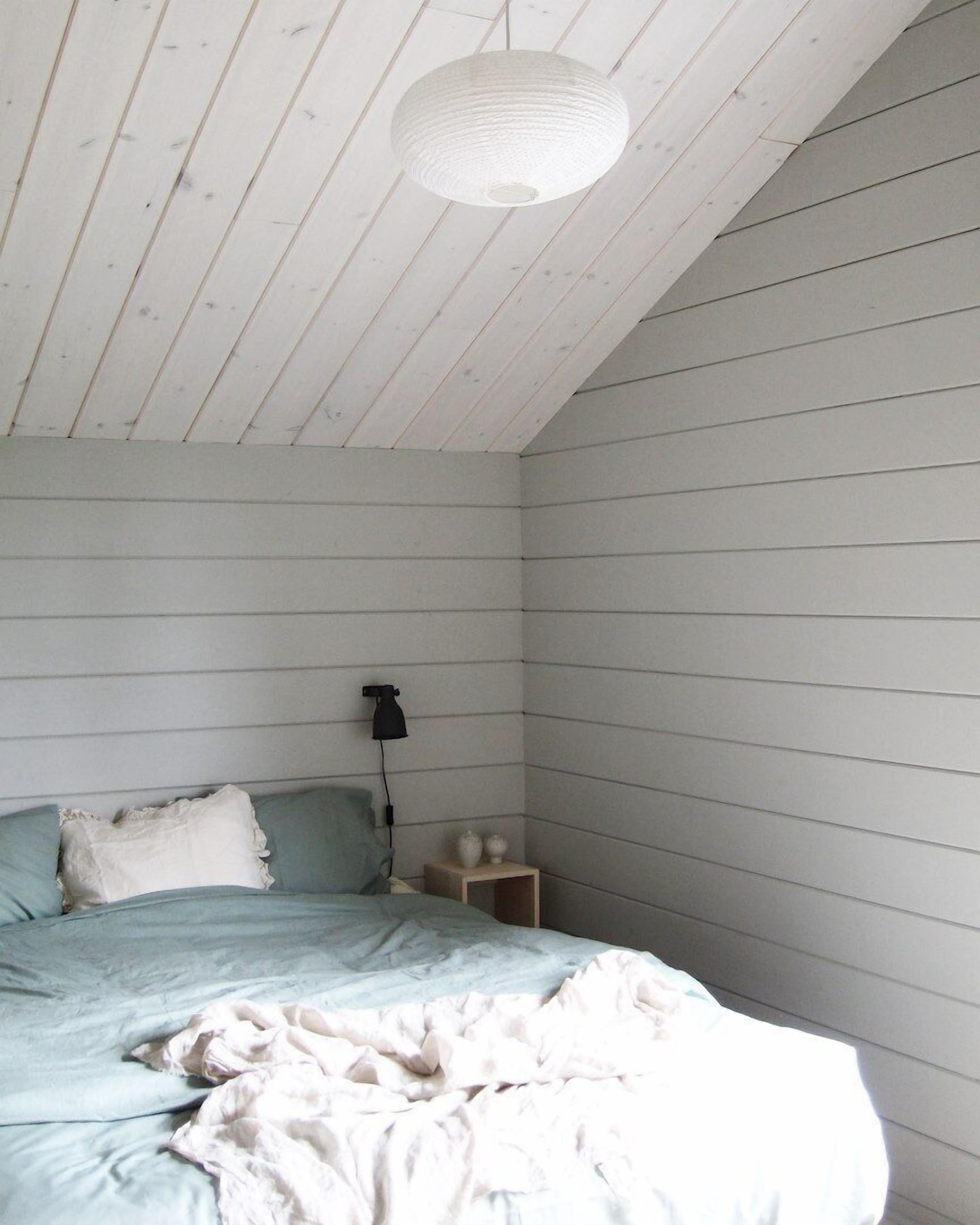 A bedroom with a sloped ceiling, a bed with a light blue bedspread, a white pillow, and a bedside table with a lamp.