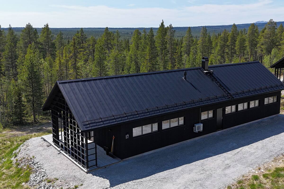 A black cabin with a sloped roof, a wooden deck, and a gravel driveway.