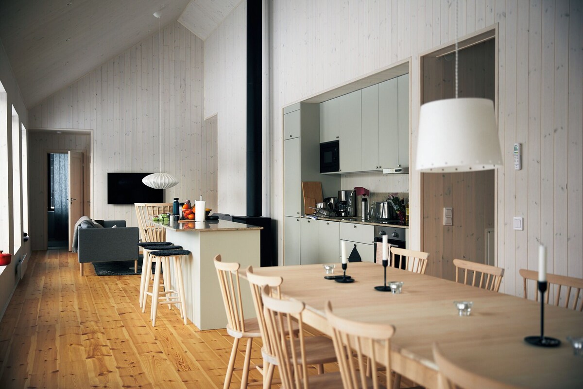 A modern kitchen with wooden cabinets, a dining area with a long table and chairs, and a TV mounted on the wall.