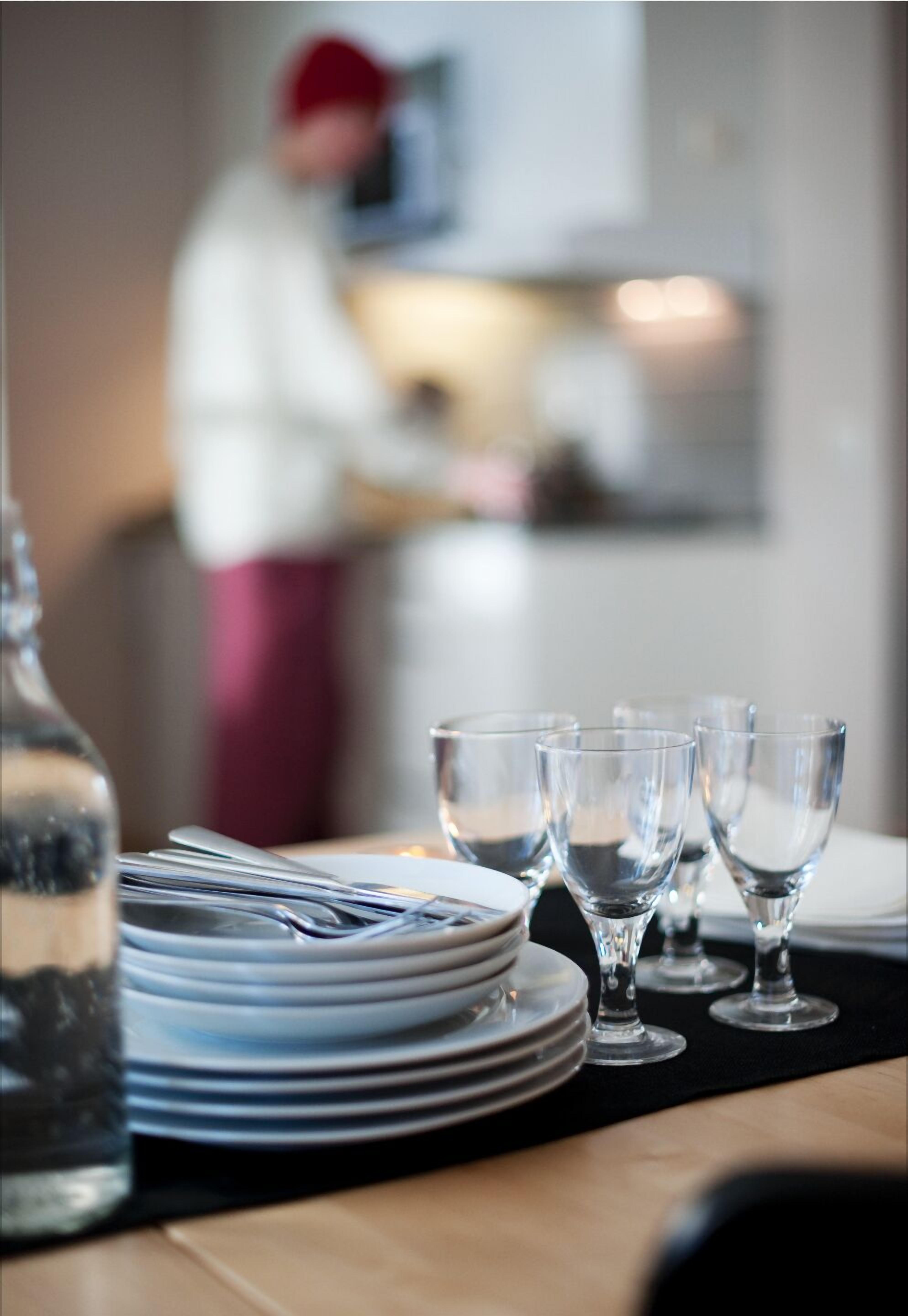 Een eettafel met gestapelde borden en heldere glazen, een fles en een vage figuur op de achtergrond.