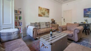 Een woonkamer met een bank, fauteuil, salontafel, boekenkast en schilderijen.