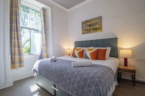 A neatly made bed with a patterned bedspread, a bedside table with a lamp, and a window with curtains. - Historic 2-Bed Paisley Retreat | Airport & Station (Paisley)
