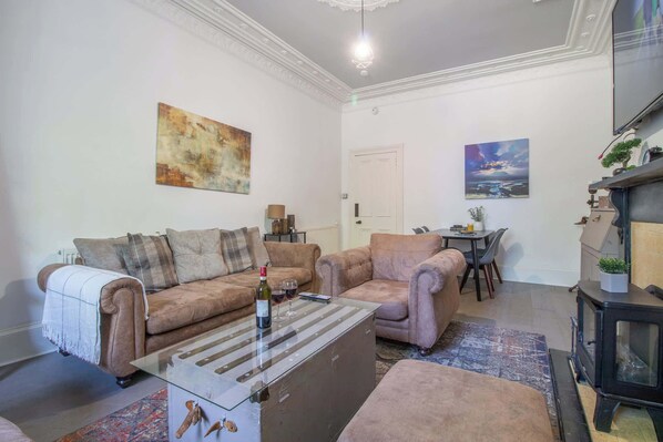 Uma sala de estar com sofá, mesa de centro de vidro com garrafa e copos, lareira e duas pinturas nas paredes.