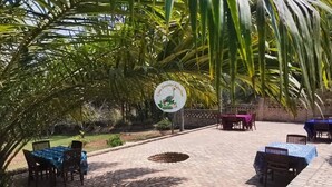 An outdoor patio area with brick flooring, covered tables, and chairs, surrounded by palm trees and greenery. - chez Drocella Eglantine Lodge Hotel (Kigali)