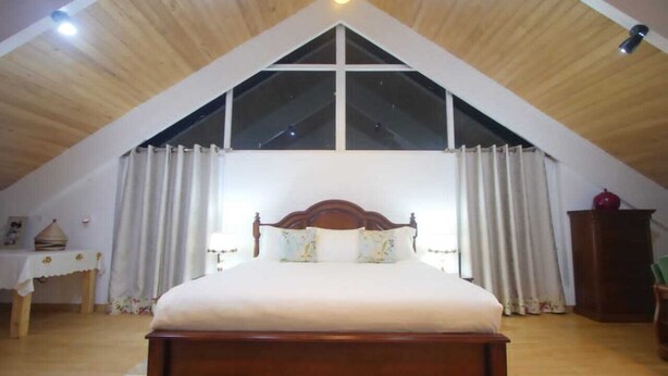 A bedroom with a wooden ceiling, a large window with curtains, a wooden bed, and a nightstand with a lamp. - chez Drocella Eglantine Lodge Hotel (Kigali)