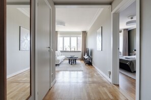 Una habitació d'hotel moderna amb sòl de fusta, un sofà blanc, una taula de dinar amb cadires, un llit amb edredó gris i una finestra gran amb cortines.