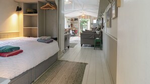 A compact bedroom with a bed, a small seating area, and a kitchenette in the background. - The Artist's Cabin By The Sea (Herne Bay)
