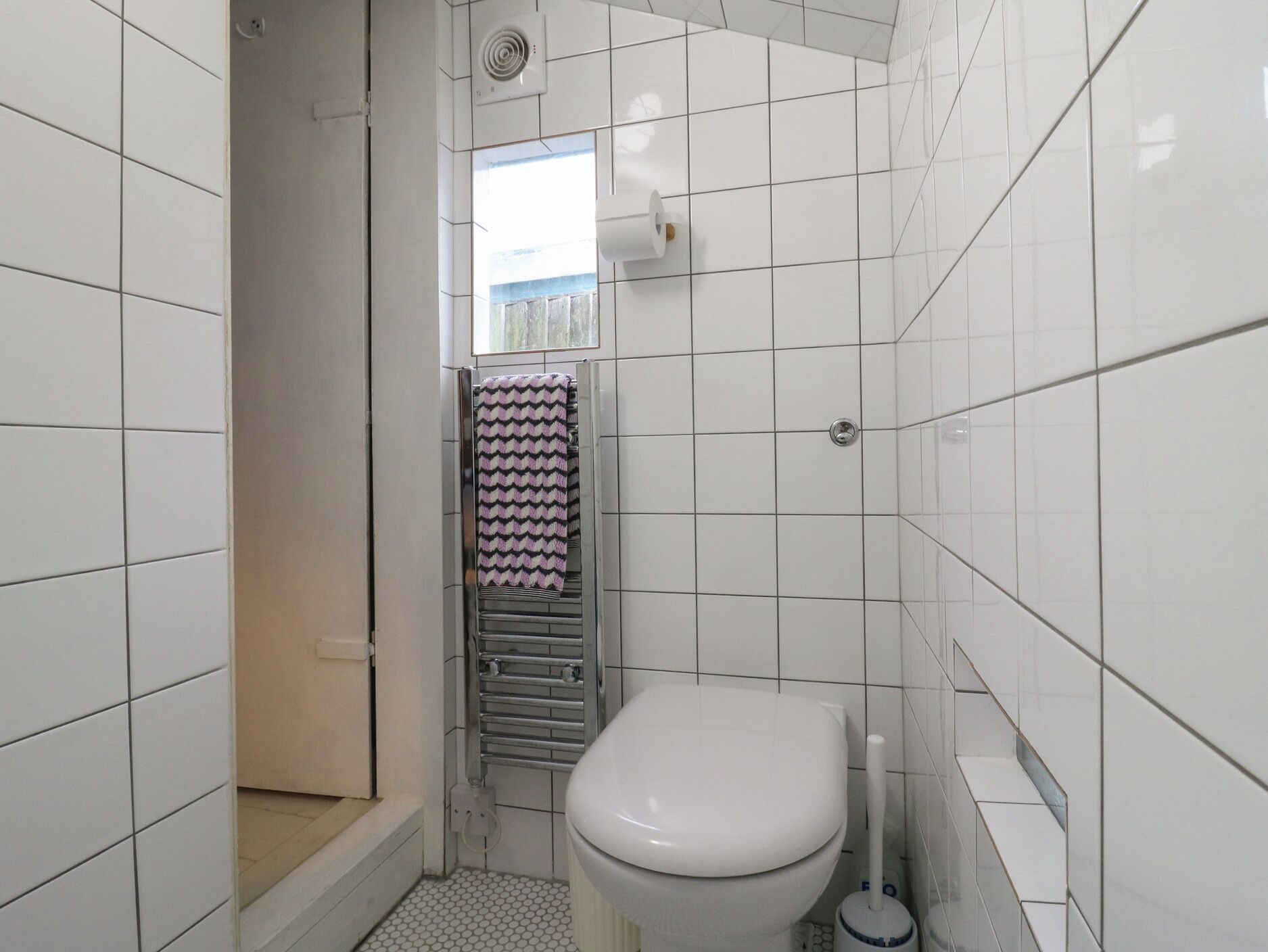 A small, tiled bathroom with a white toilet, a towel rack with a patterned towel, and a visible shower area.