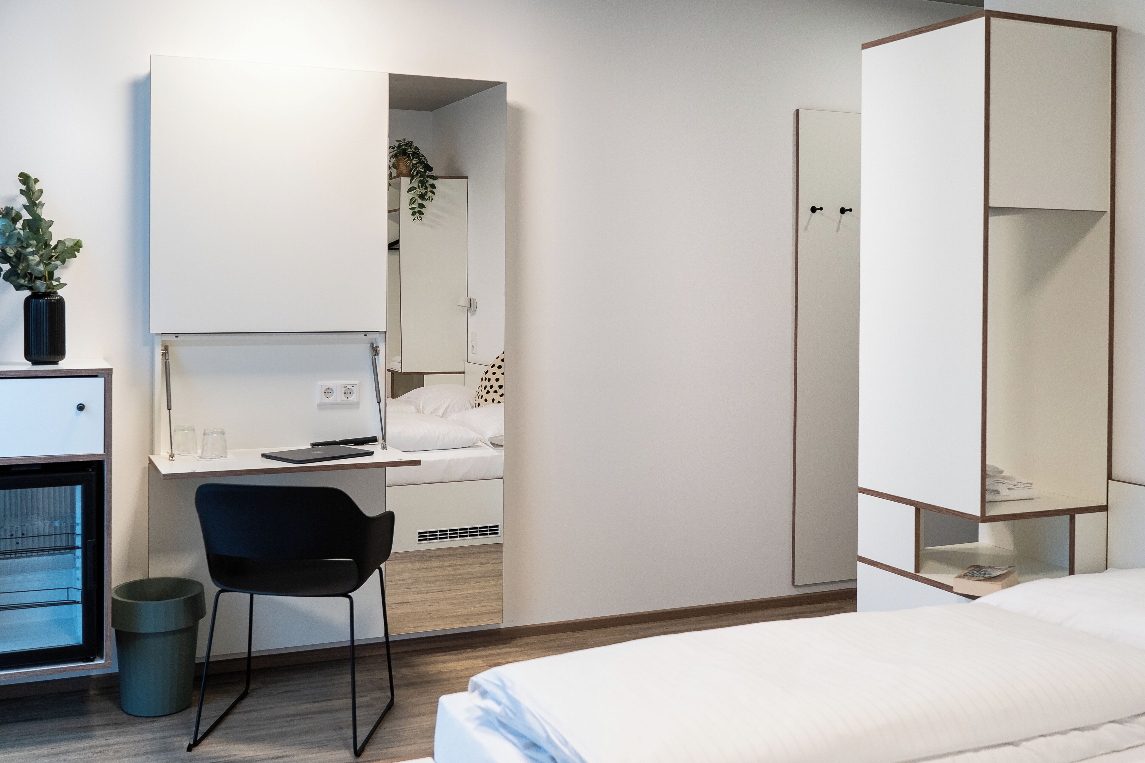 Ett modernt hotellrum med en säng, ett skrivbord med en stol, en garderob och en papperskorg.