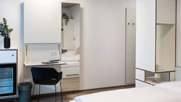 Ett modernt hotellrum med en säng, ett skrivbord med en stol, en garderob och en papperskorg.