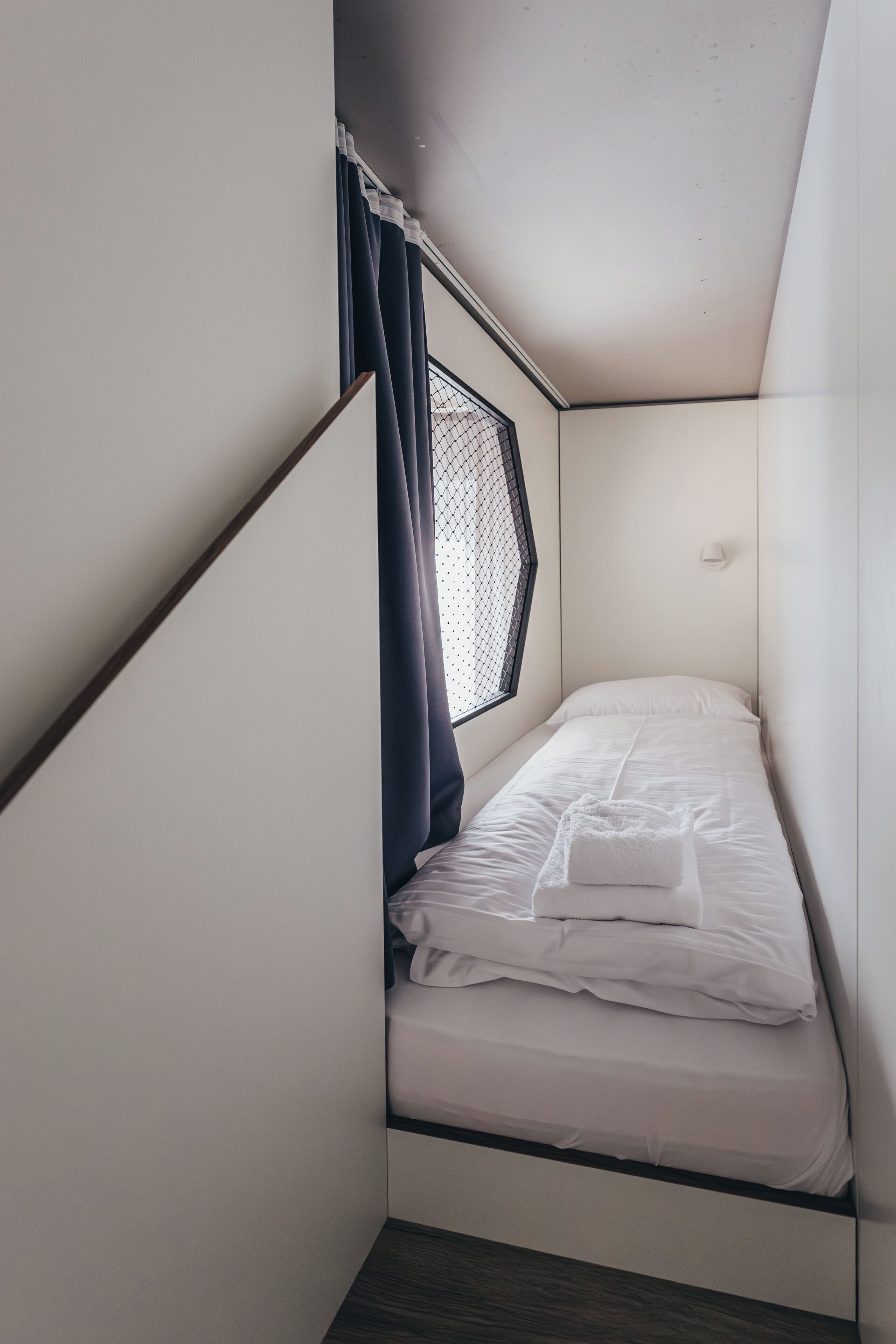 A small, compact bedroom with a single bed, a window with curtains, and a white wall.