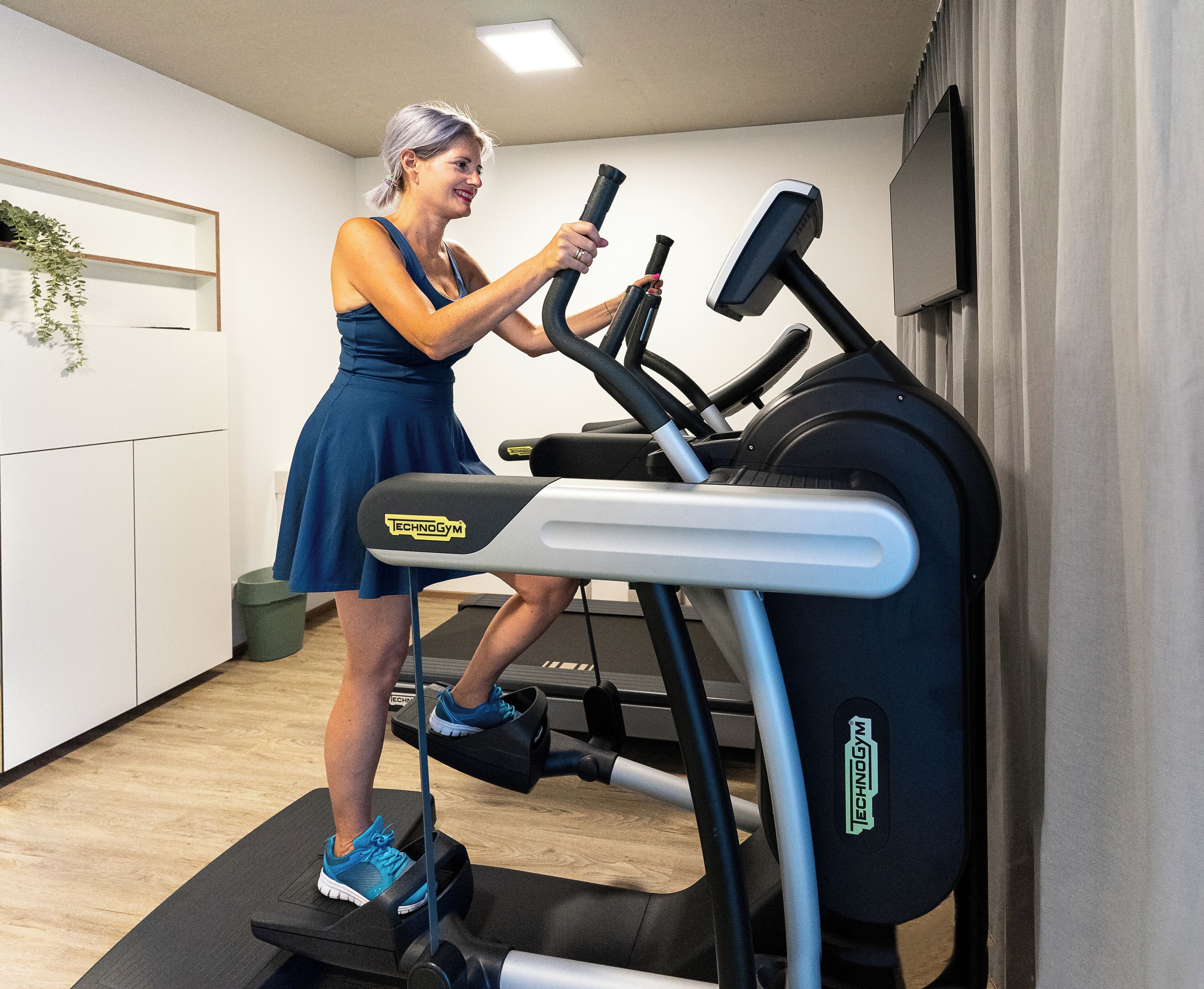 A woman using a treadmill in a room with a white wall, a small plant, and a wooden floor.
