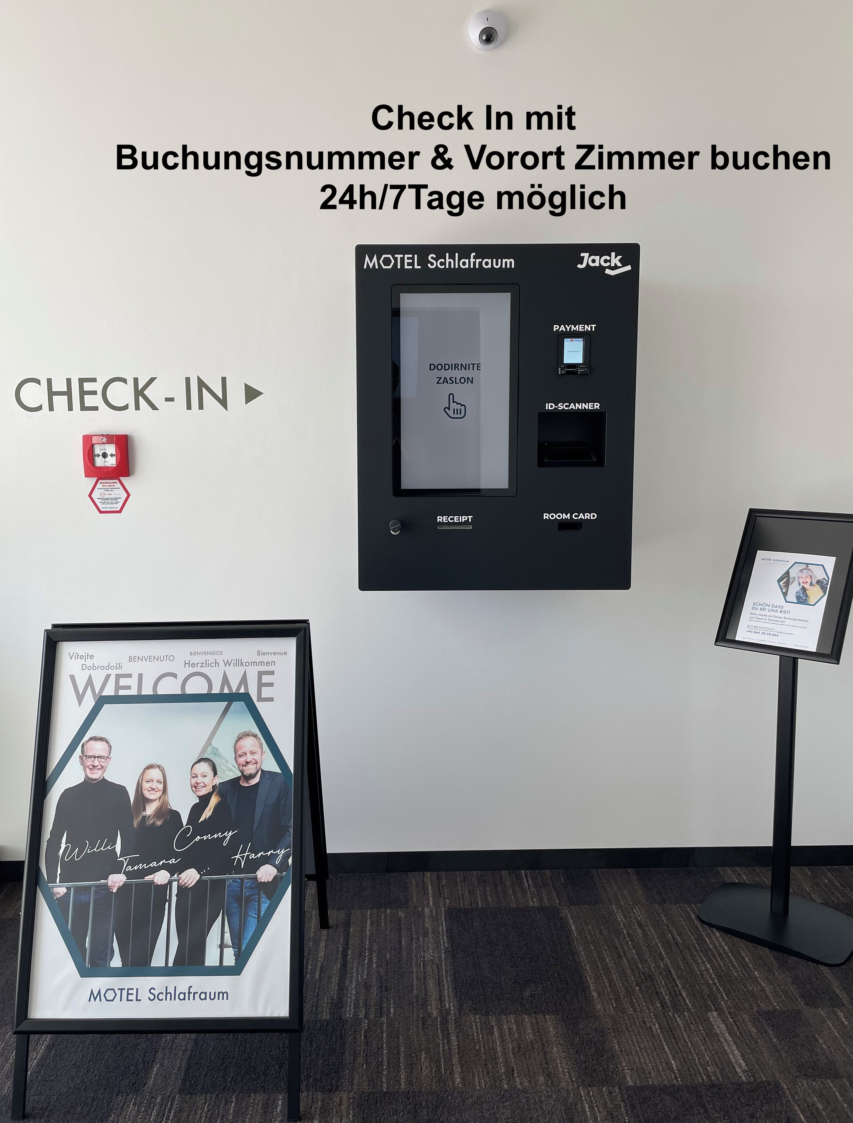 A check-in station with a digital display for room selection, a payment terminal, and a framed welcome sign.