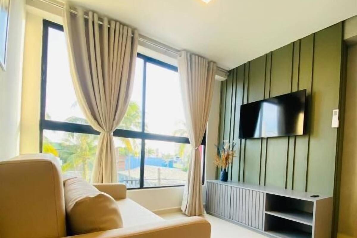 Una sala de estar moderna con un sofá, un televisor de pantalla plana montado en una pared verde y un ventanal con cortinas.