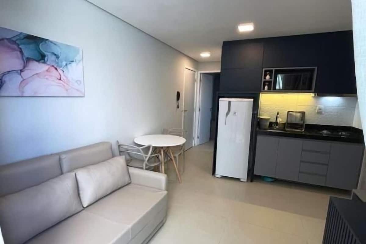 Una sala de estar moderna con un sofá, una pequeña mesa redonda y una zona de cocina con armarios y un refrigerador.