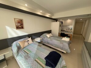 A modern hotel room with two beds, a small desk, and a kitchenette.