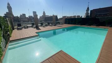 Una piscina en la azotea con terraza de madera y sillas reclinables, rodeada de edificios de la ciudad y un cielo despejado.