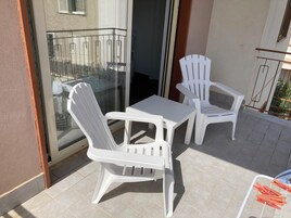 Varanda com duas cadeiras de plástico brancas, uma mesa pequena, uma porta de vidro e guarda-corpo de metal.