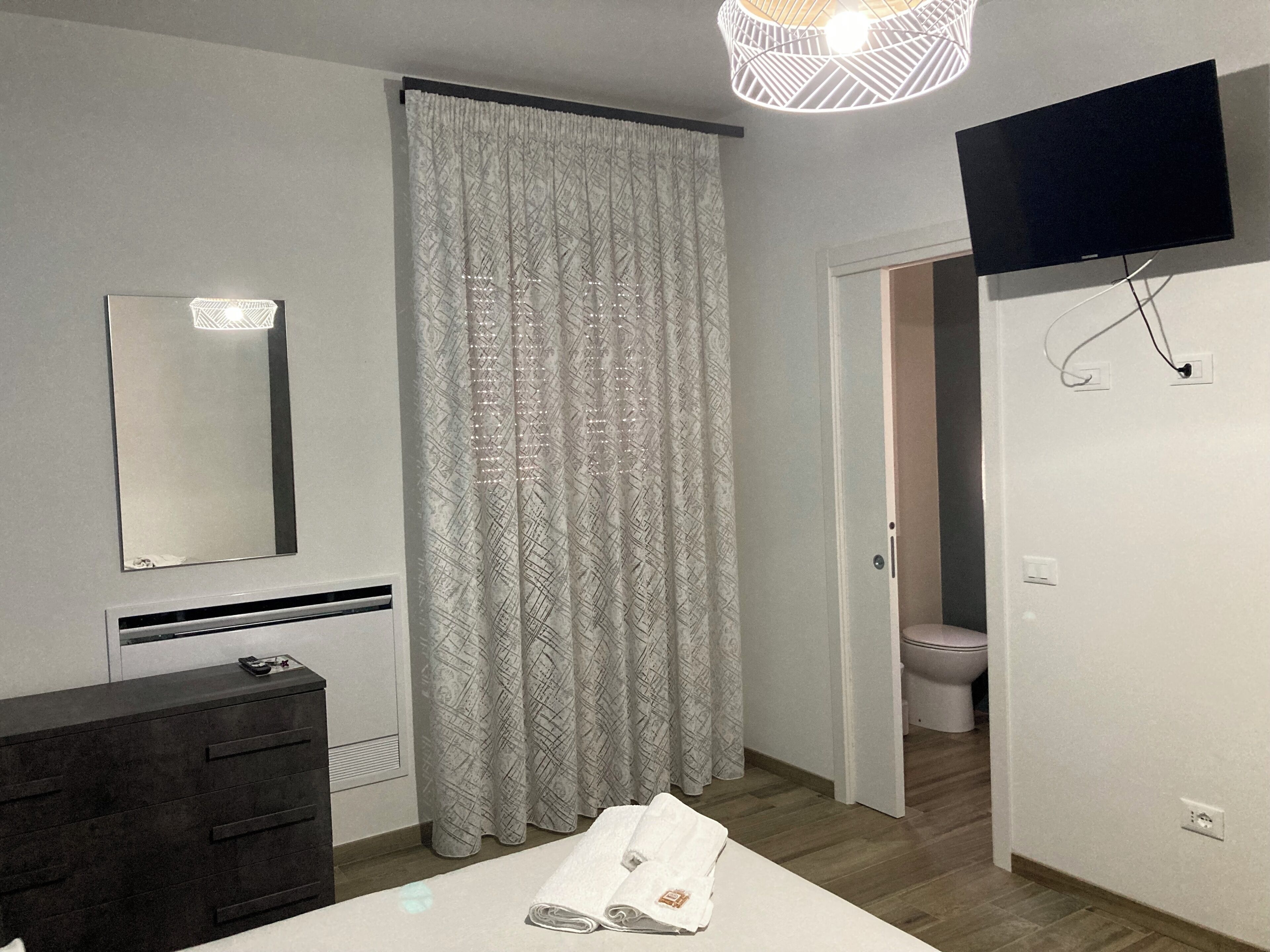 Ein modernes Schlafzimmer mit einem großen Bett, einem Nachttisch, einem Spiegel, einem an der Wand montierten Fernseher und einem durch eine offene Tür einsehbaren Badezimmer.