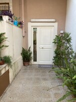 Um pátio estreito com uma porta branca, plantas em vasos e piso de azulejos.