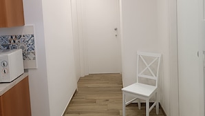 Een smalle gang met witte wanden, houten vloer en een witte stoel. Links staan kastjes en een magnetron, en aan het einde een deur.