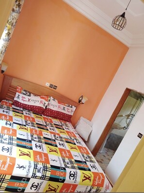 Une chambre à coucher avec un lit recouvert d’une couette à motifs, une tête de lit en bois, un luminaire fixé au mur et une porte donnant sur une salle de bain.