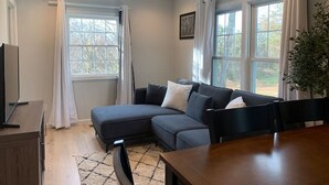 Een moderne woonkamer met een hoekbank, een houten eettafel en grote ramen met gordijnen.