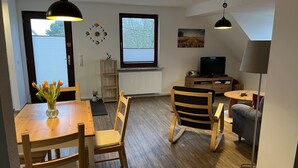 A modern living room with a wooden dining table, chairs, a TV, and a sofa. - Urlaub auf dem Bauernhof in der Nähe von Sankt Peter Ording (Tating)