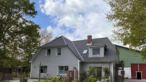 A two-story house with a grey exterior, a visible chimney, and a small attached garage. - Urlaub auf dem Bauernhof in der Nähe von Sankt Peter Ording (Tating)