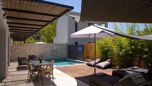 Un área junto a la piscina con mesa y sillas, un espacio de descanso con cojines y un porche cubierto.