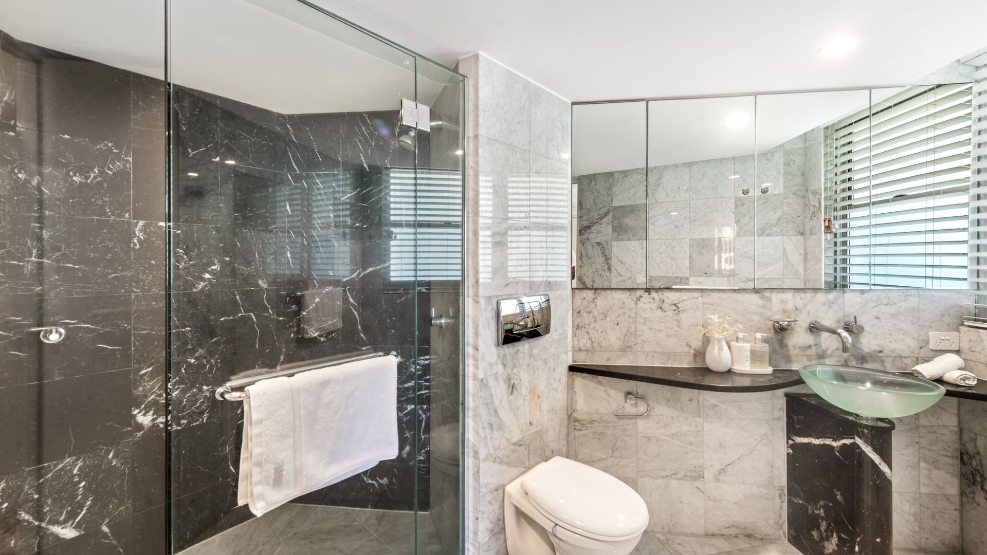 A modern bathroom with a glass-enclosed shower, a marble-tiled wall, a white toilet, and a glass sink.