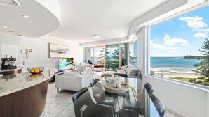Una sala moderna con un ventanal grande que da a una playa, una zona de cocina con cafetera y un comedor con mesa de vidrio.