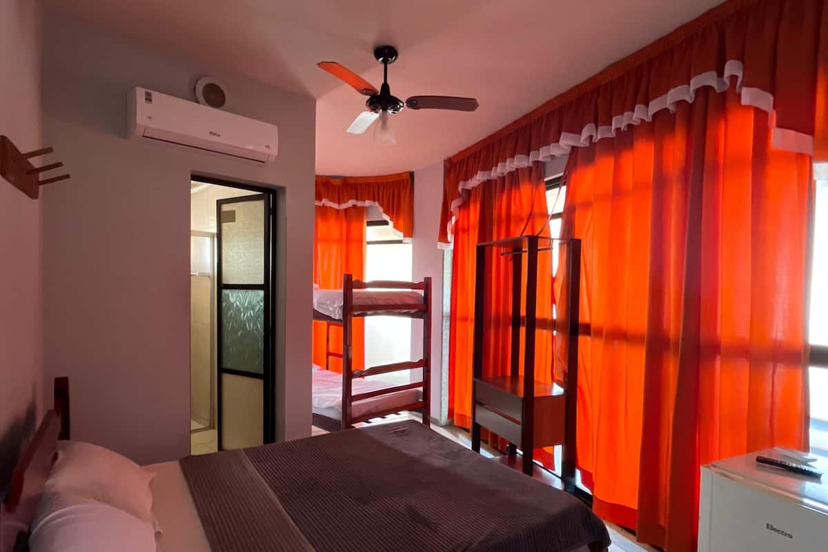 Un dormitorio con cama, ventilador de techo, equipo de aire acondicionado, escritorio con silla y ventana con cortinas naranjas.
