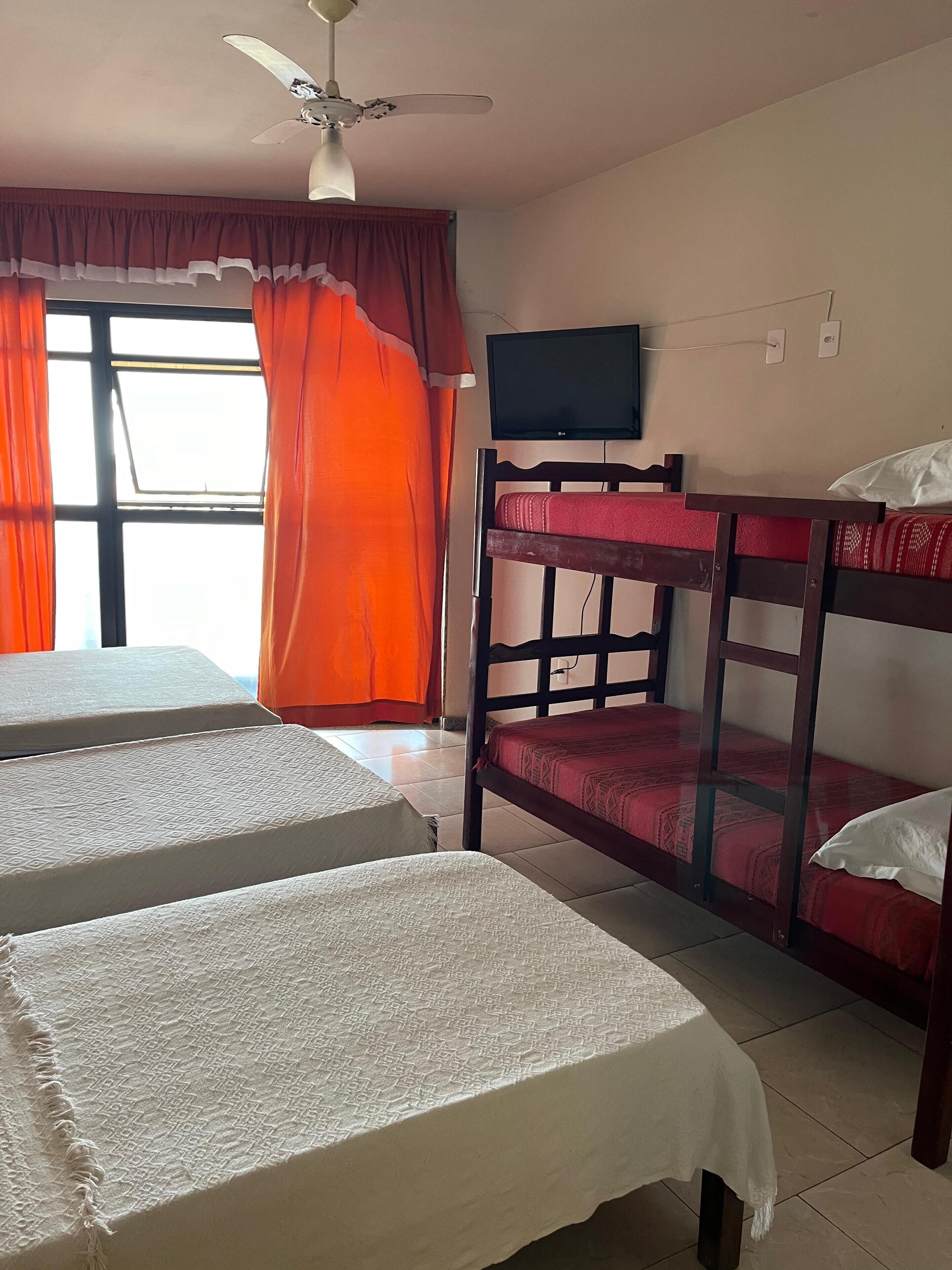 A dormitory room with bunk beds, a ceiling fan, a TV, and a window with curtains.