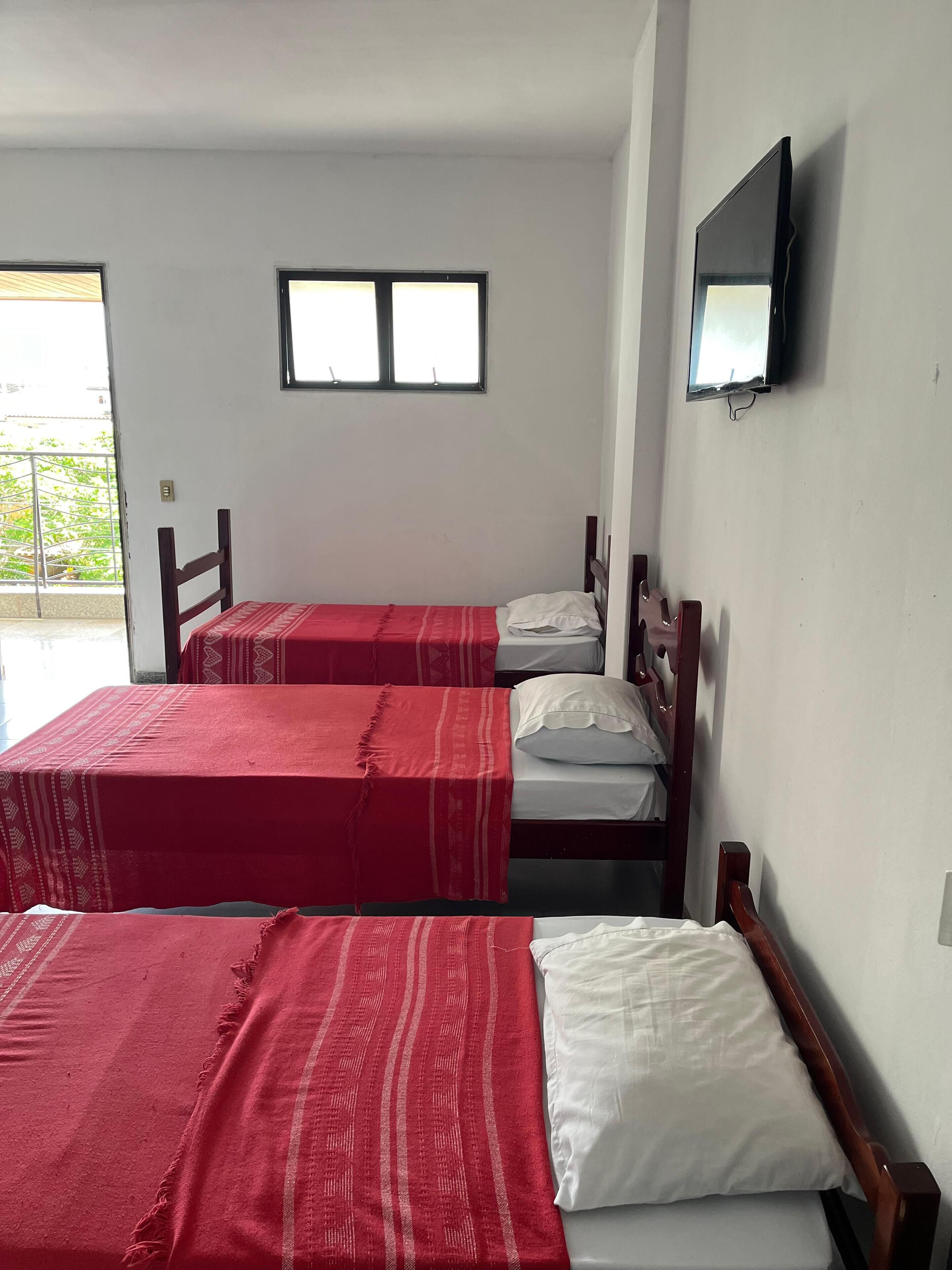 A room with three single beds, each with a wooden headboard and a red bedspread. There is a television mounted on the wall to the right.