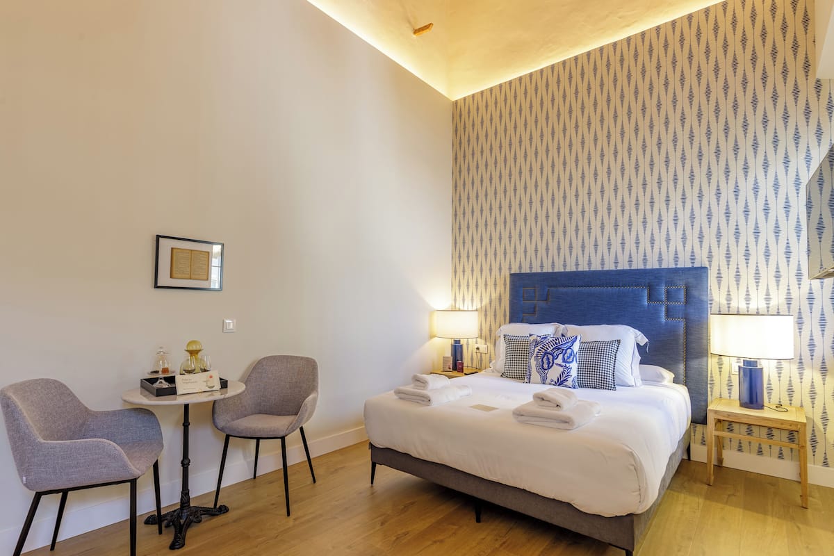 Una habitación de hotel con cama, dos sillas, una mesita y una mesita de noche con lámparas.