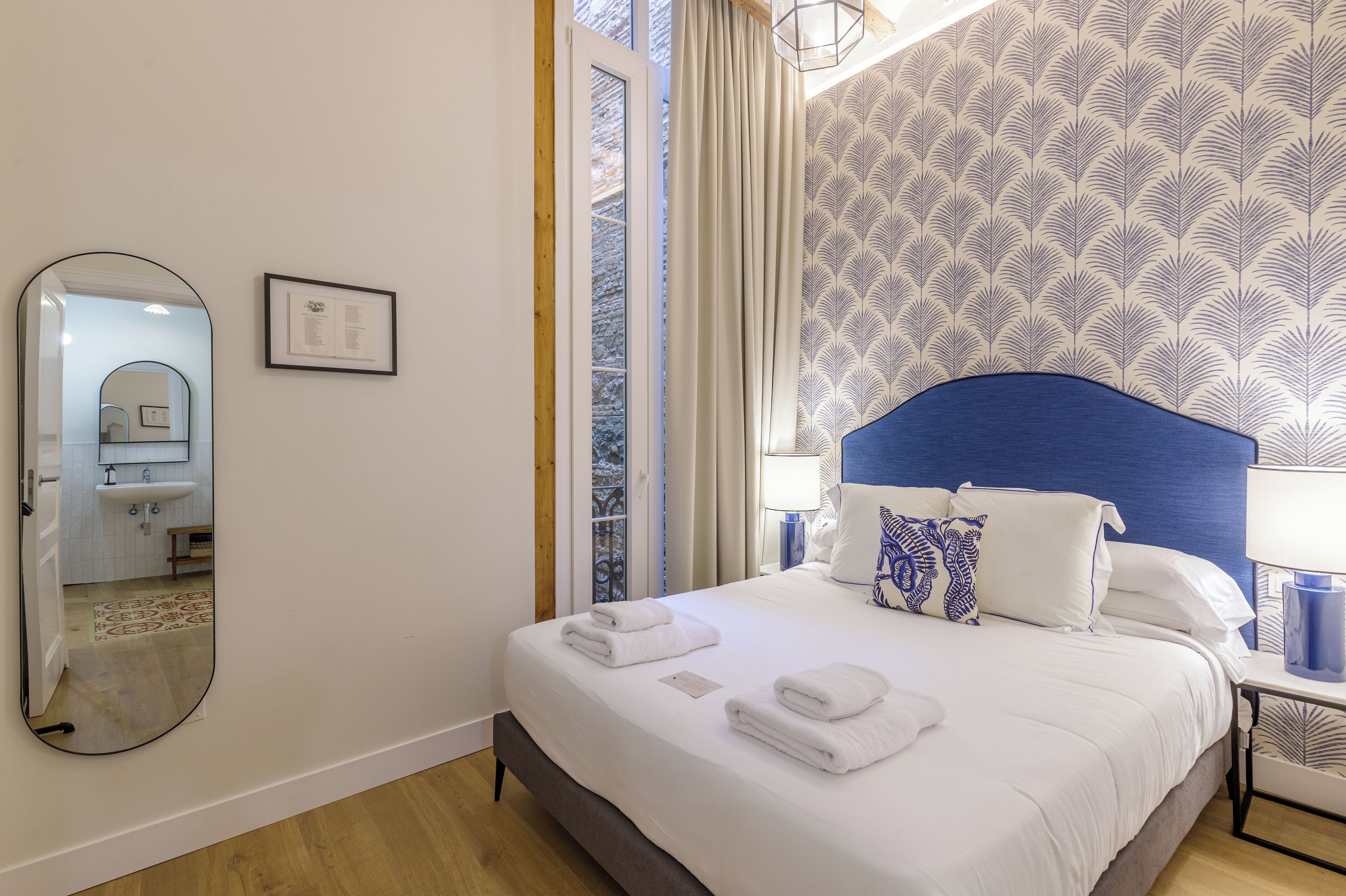 A neatly made bed with white linens and towels, a blue headboard, and a patterned wallpaper.