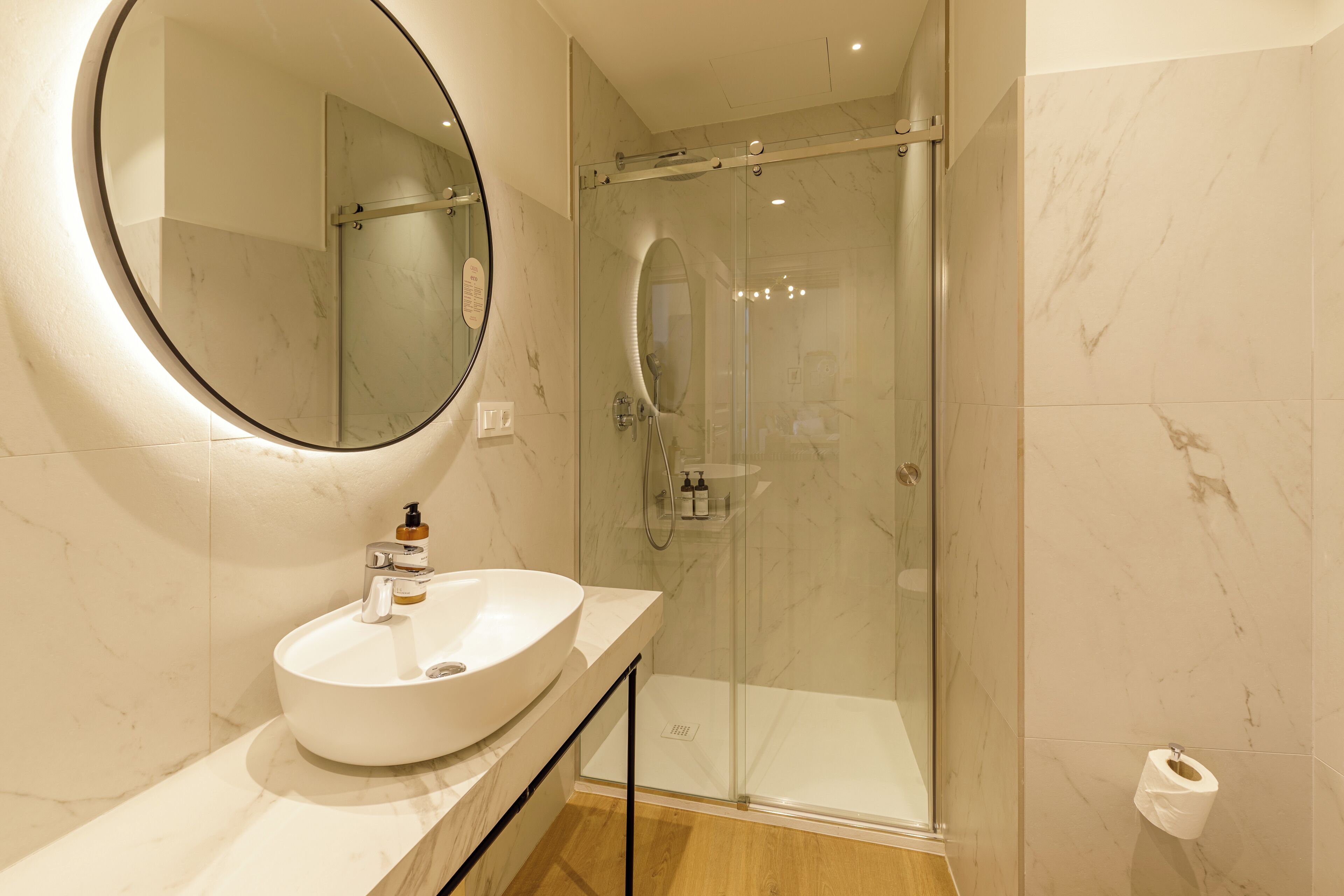 A modern bathroom with a round mirror, a white sink, and a glass-enclosed shower.
