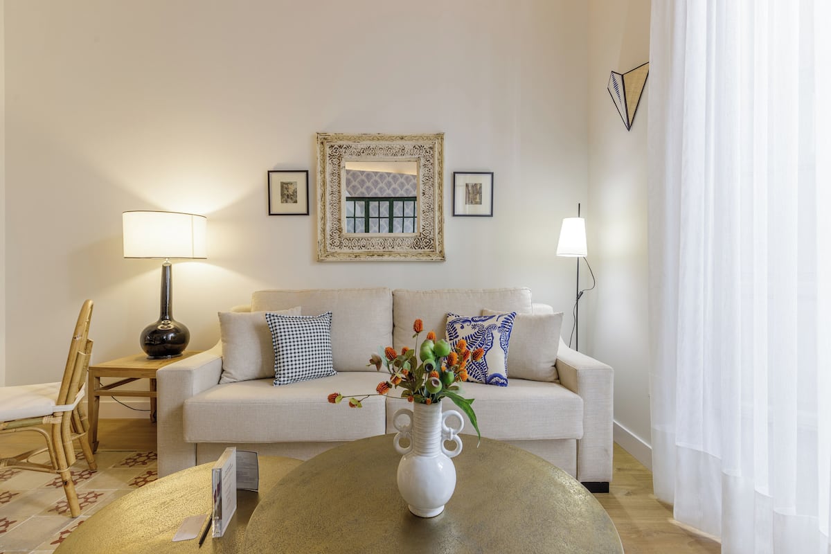 Una sala de estar con un sofá, dos lámparas, un jarrón decorativo con flores y una alfombra estampada.