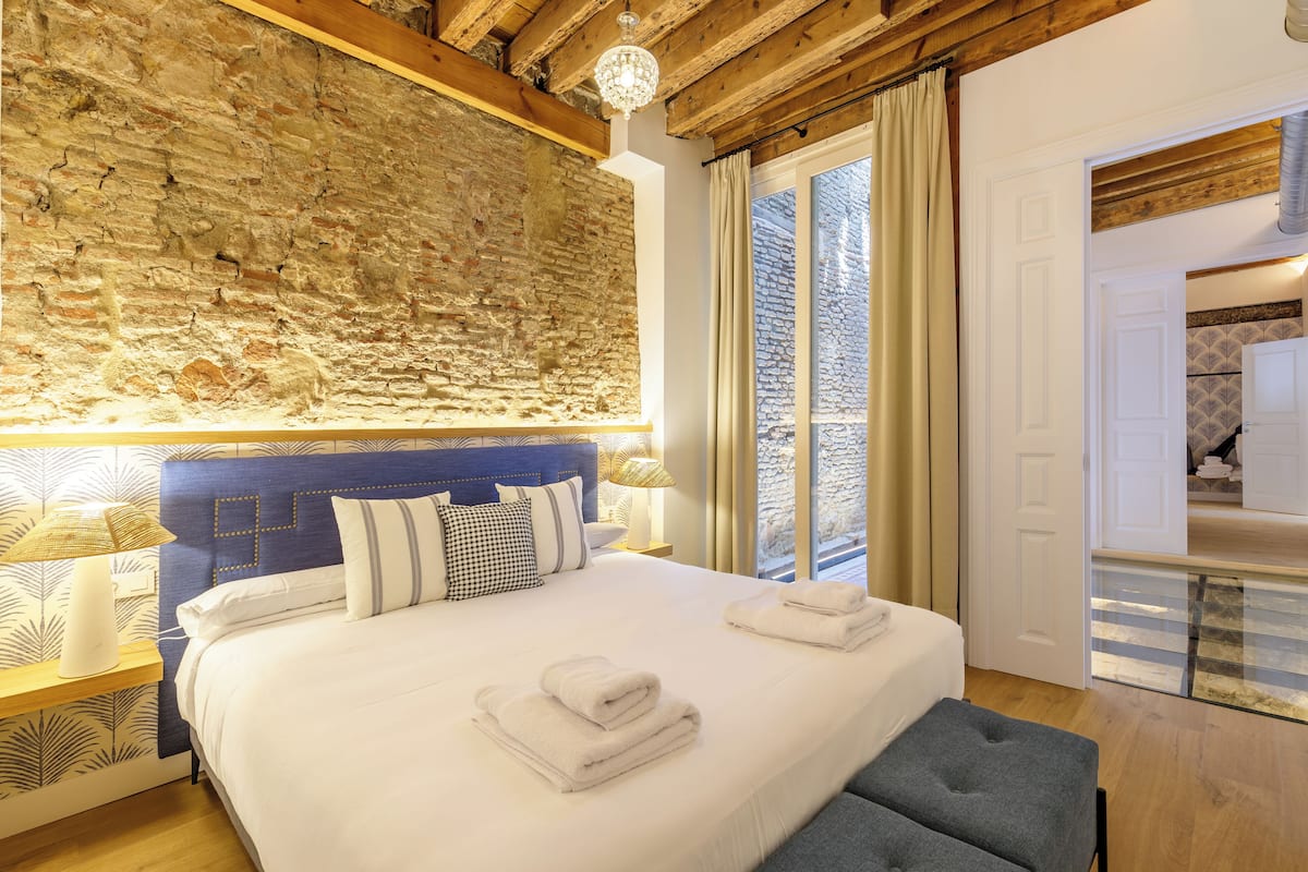 Un dormitorio con pared de piedra, una cama con ropa de cama blanca, un cabecero azul y una puerta de vidrio que da al baño.