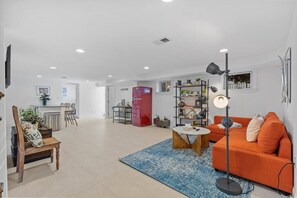 Un vaste espace de vie comprenant un canapé, une table basse et un lampadaire. Au fond, on aperçoit un coin cuisine avec un réfrigérateur et une table à manger.