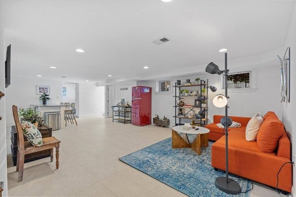 A spacious living area with a sofa, a coffee table, and a floor lamp. There is a kitchen area in the background with a refrigerator and a dining table.